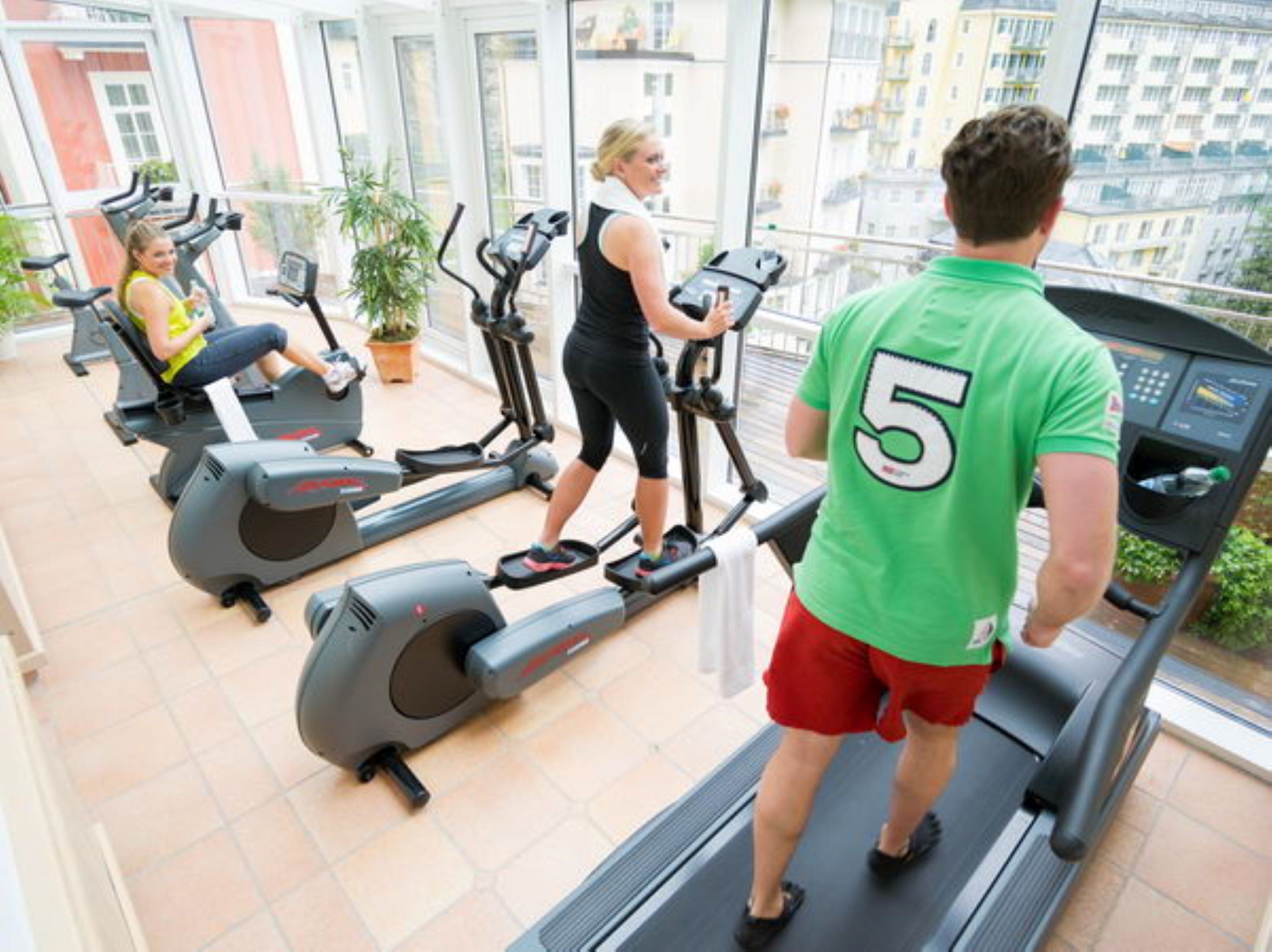 A gym with treadmills and exercise bikes, large windows, and a view of buildings.