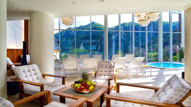 A modern lounge area with white cushioned chairs, a wooden table with a fruit bowl, and large windows offering a view of a mountain and a swimming pool. - Villa Orania (Bad Gastein)