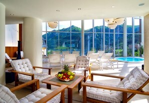 Un espacio lounge moderno con sillas blancas acolchadas, una mesa de madera con frutero, y grandes ventanales con vistas a una montaña y una piscina.