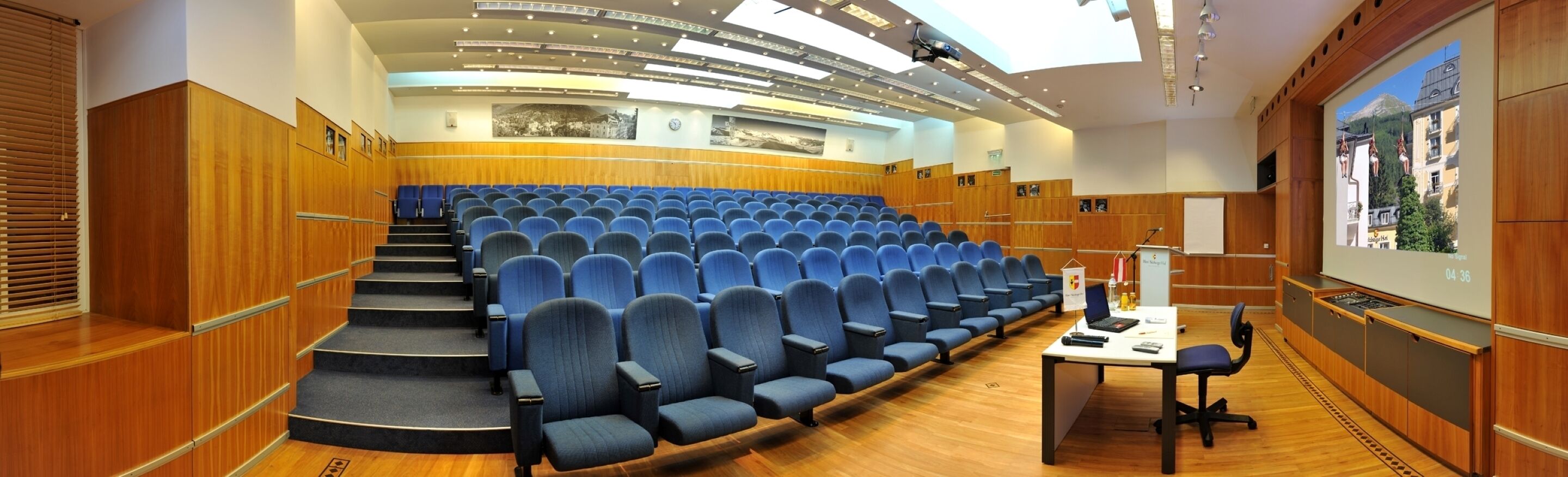 A lecture hall with rows of blue chairs, wooden paneling, a projector screen, and a small table with a laptop.