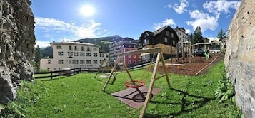 Um parquinho com balanços e uma cesta de basquete, cercado por edifícios e uma montanha ao fundo.