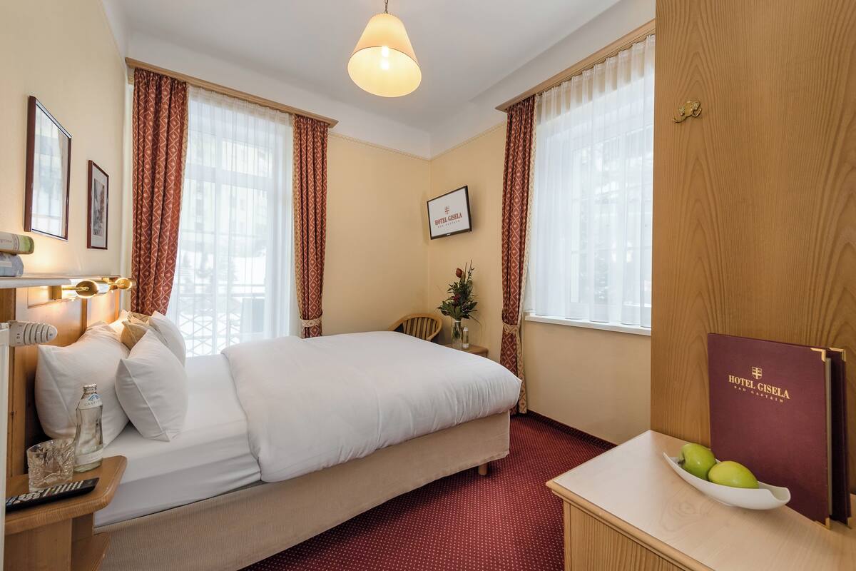 A hotel room with a bed, a desk with a lamp, a chair, a vase with flowers, and a fruit bowl on the side table.