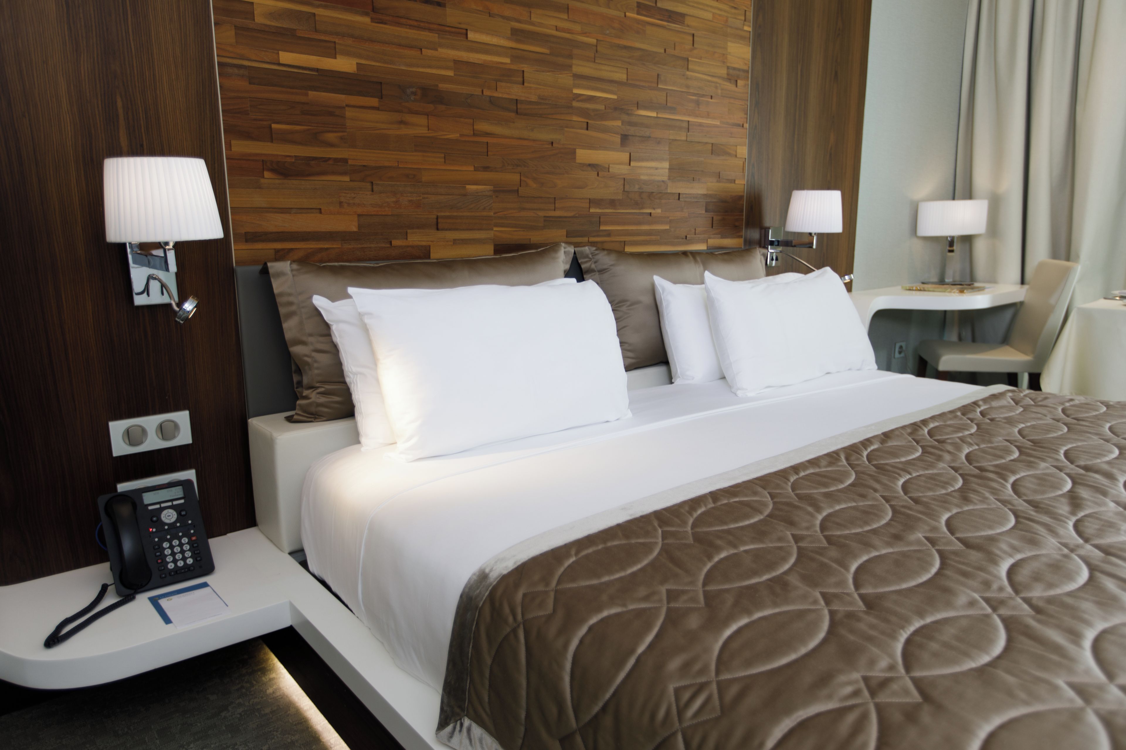 Quarto de hotel com cama, abajures, escrivaninha com telefone e luminária na parede.