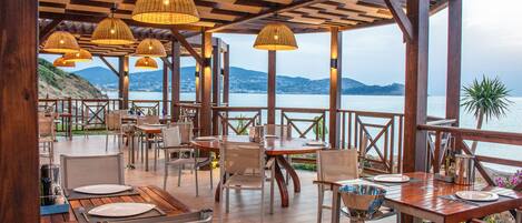 An outdoor dining area with wooden tables and chairs, wicker lamps, and a view of the sea.