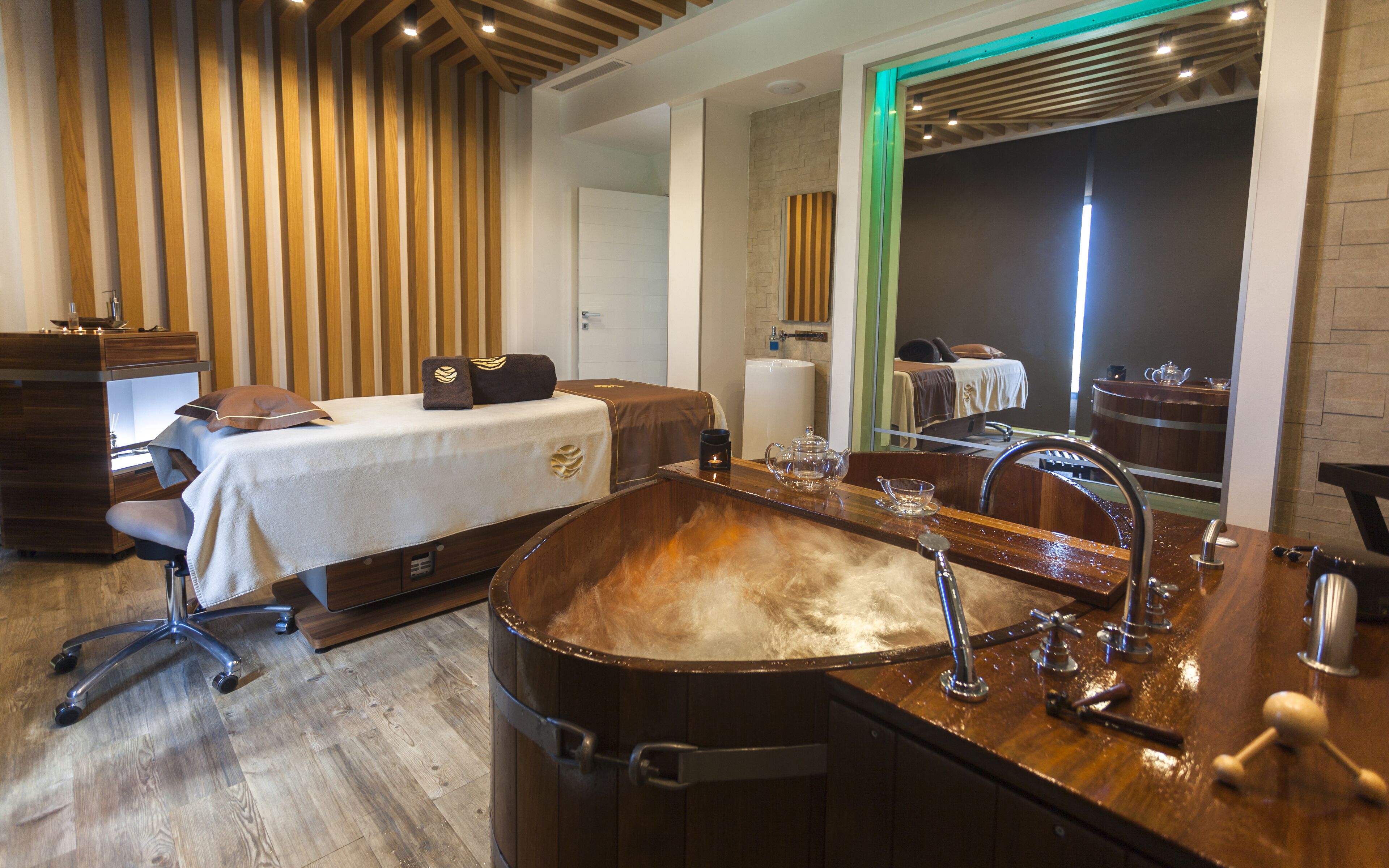 A modern spa room with a wooden slatted ceiling, a large wooden bathtub, a massage table, and a large mirror.
