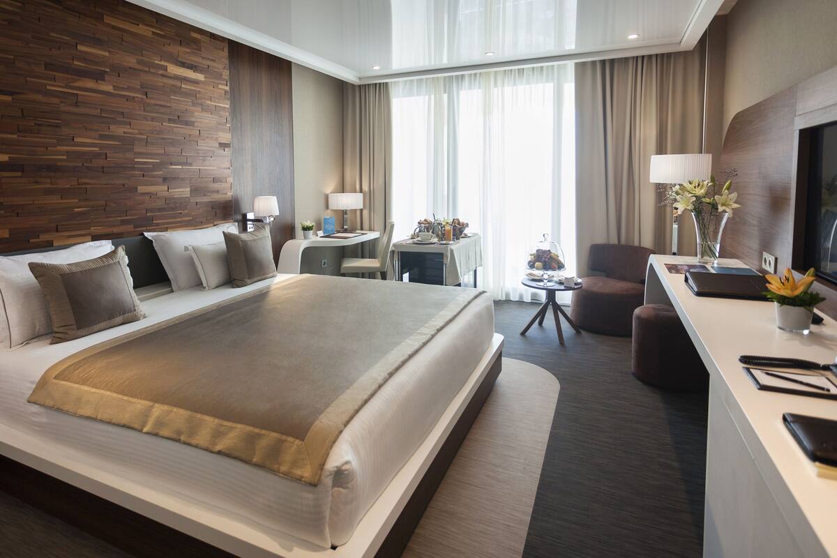 Et moderne hotelværelse med en stor seng, et skrivebord med et fjernsyn, en stol og et lille bord med lampe og blomster.