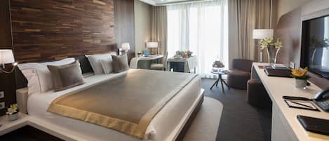 A modern hotel room with a large bed, a desk with a TV, a chair, and a small table with a lamp and flowers.