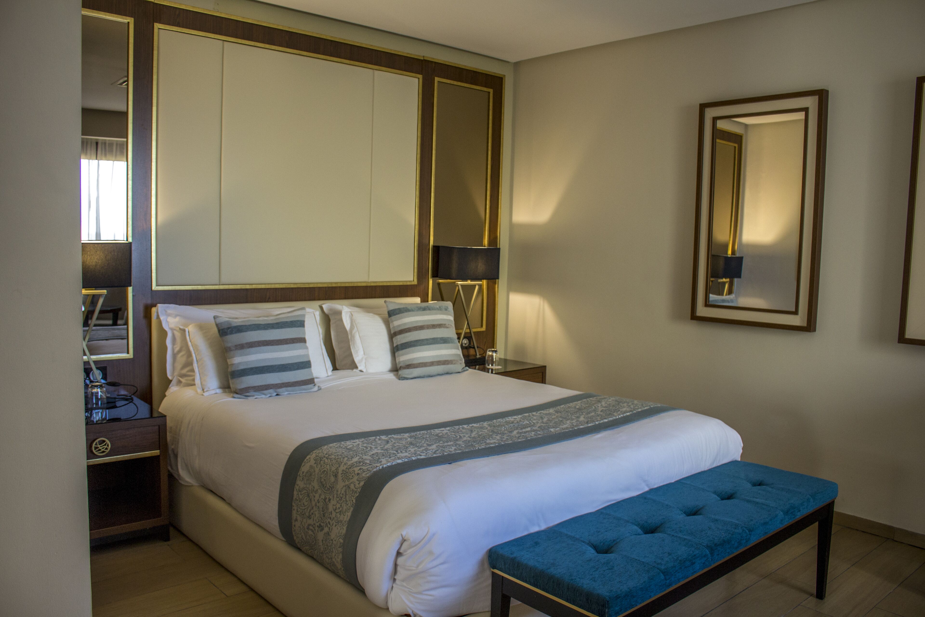 Una habitación de hotel con una cama grande, mesitas de noche, un banco y un espejo.