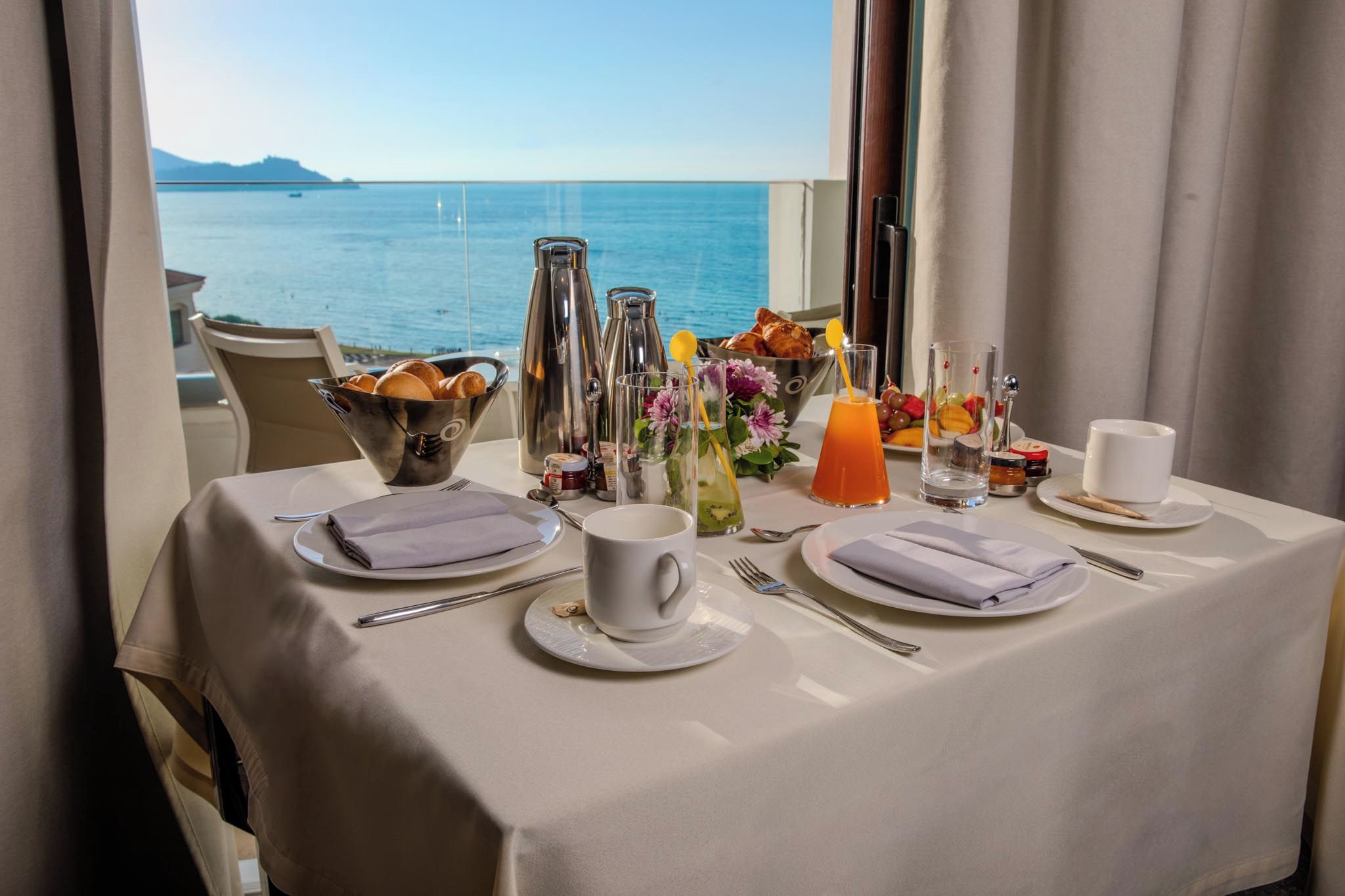 A table set for breakfast with a view of the sea, including a bowl of bread, a coffee pot, and various condiments.