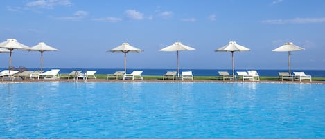 A pool with white lounge chairs and umbrellas, a clear blue sky, and a view of the ocean.