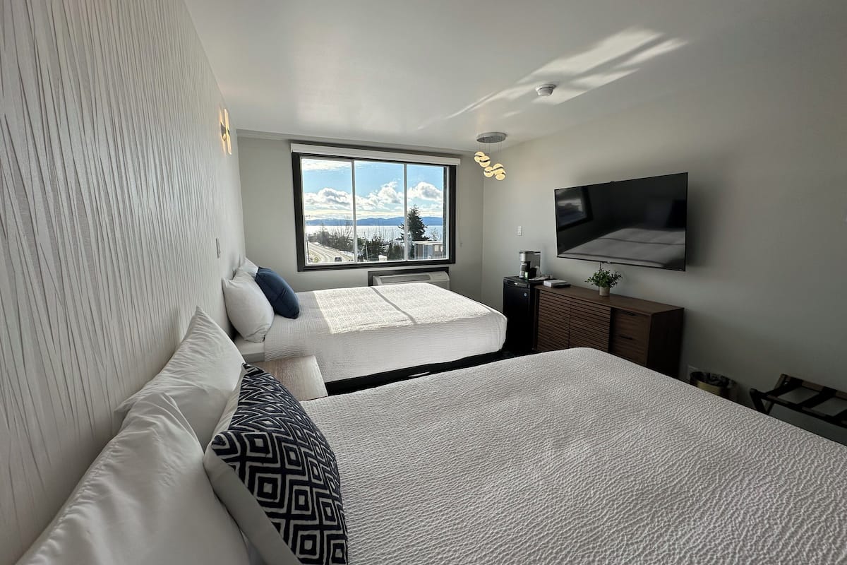 Una habitación de hotel moderna con dos camas, un ventanal con vistas, un televisor de pantalla plana y un pequeño escritorio con cafetera.