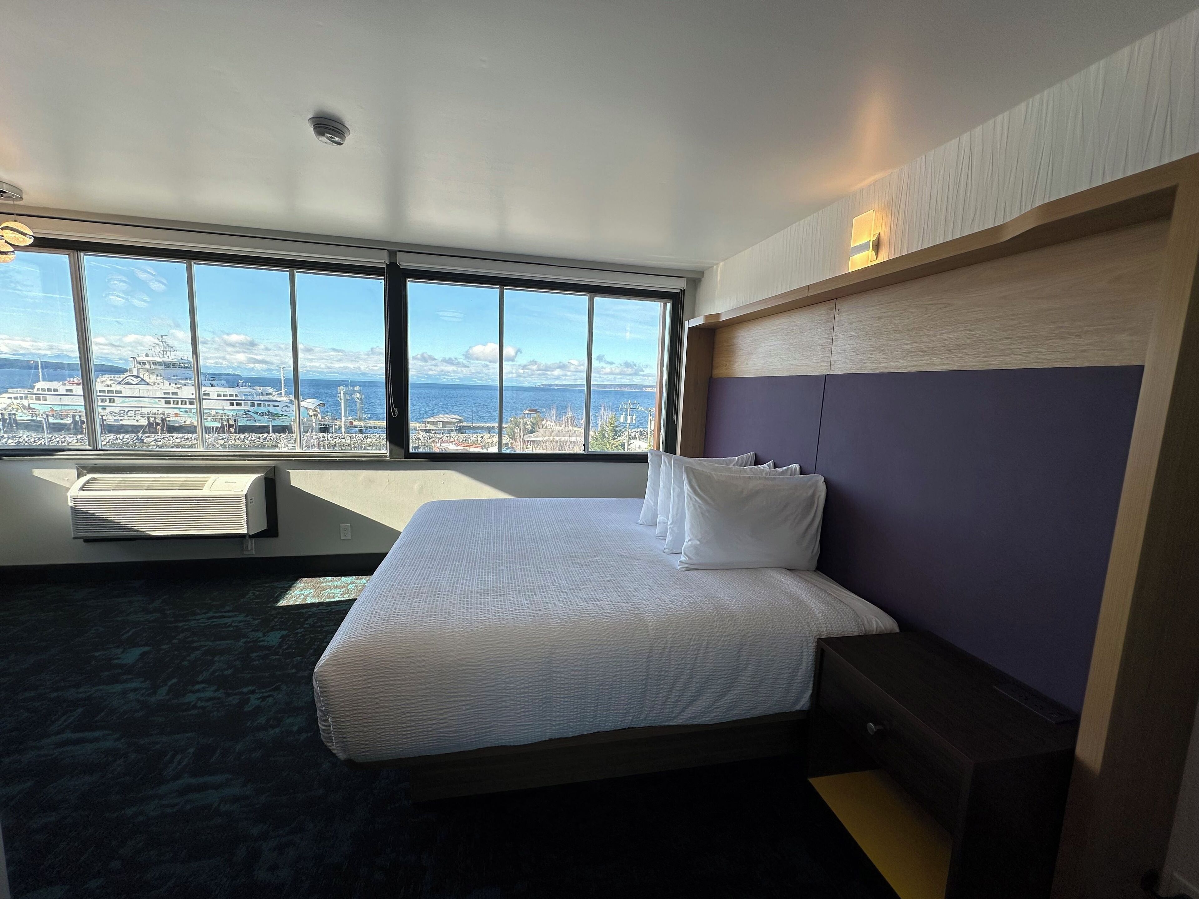 A hotel room with a large window offering a city view, a bed with white bedding, and a wooden headboard.