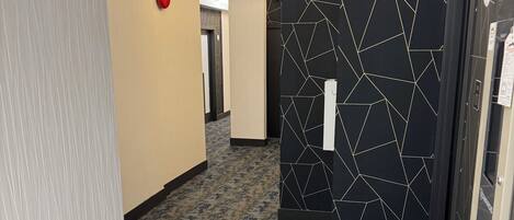 Un pasillo moderno de hotel con paredes de diseño geométrico, suelo alfombrado y un botón de emergencia rojo.