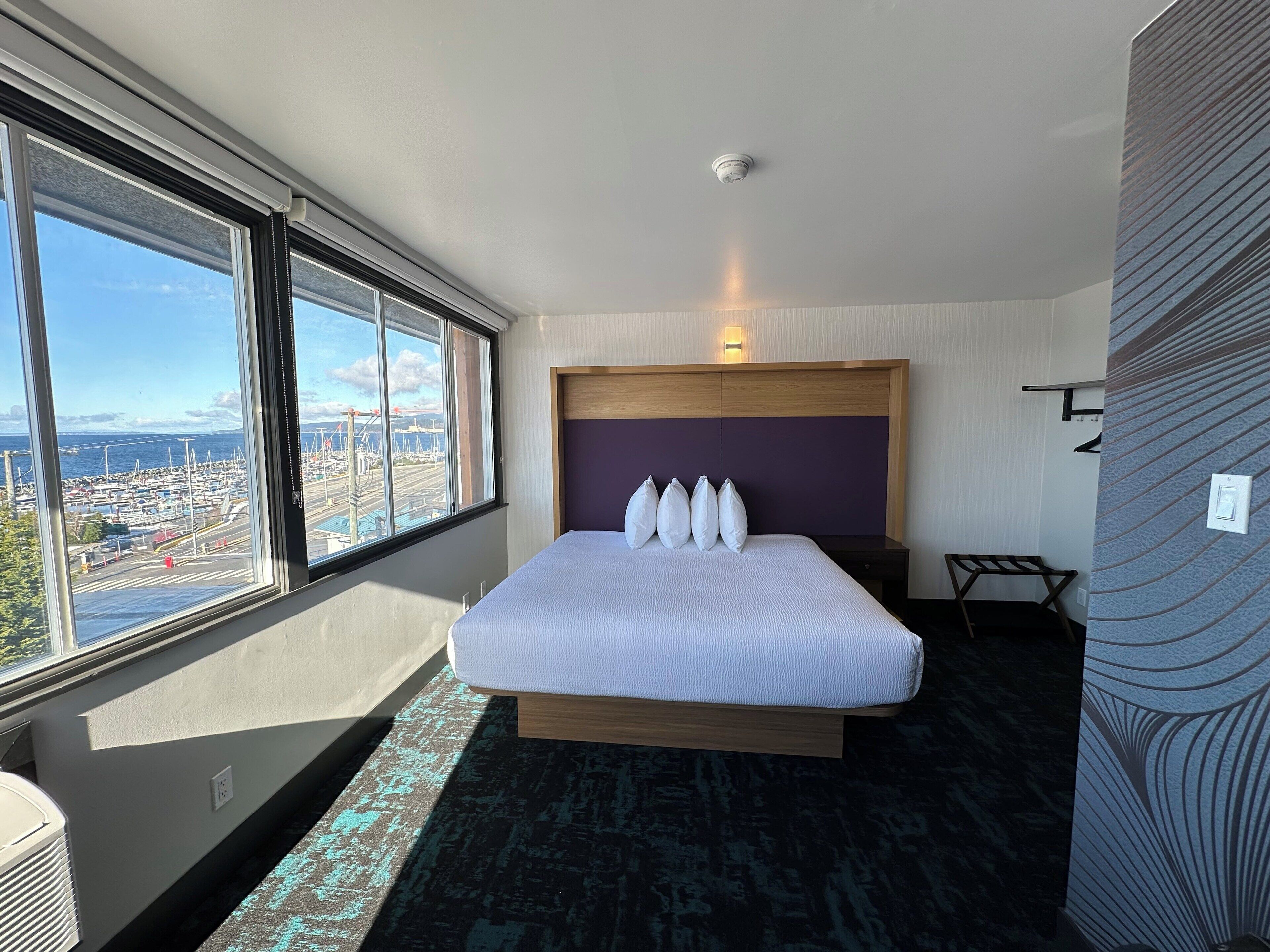 A hotel room with a large window, a bed with white bedding, a headboard, and a view of a marina.