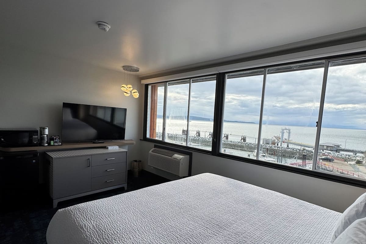 Habitación de hotel con una cama grande, un escritorio, un televisor de pantalla plana, un calefactor y vistas al mar y a estructuras industriales.