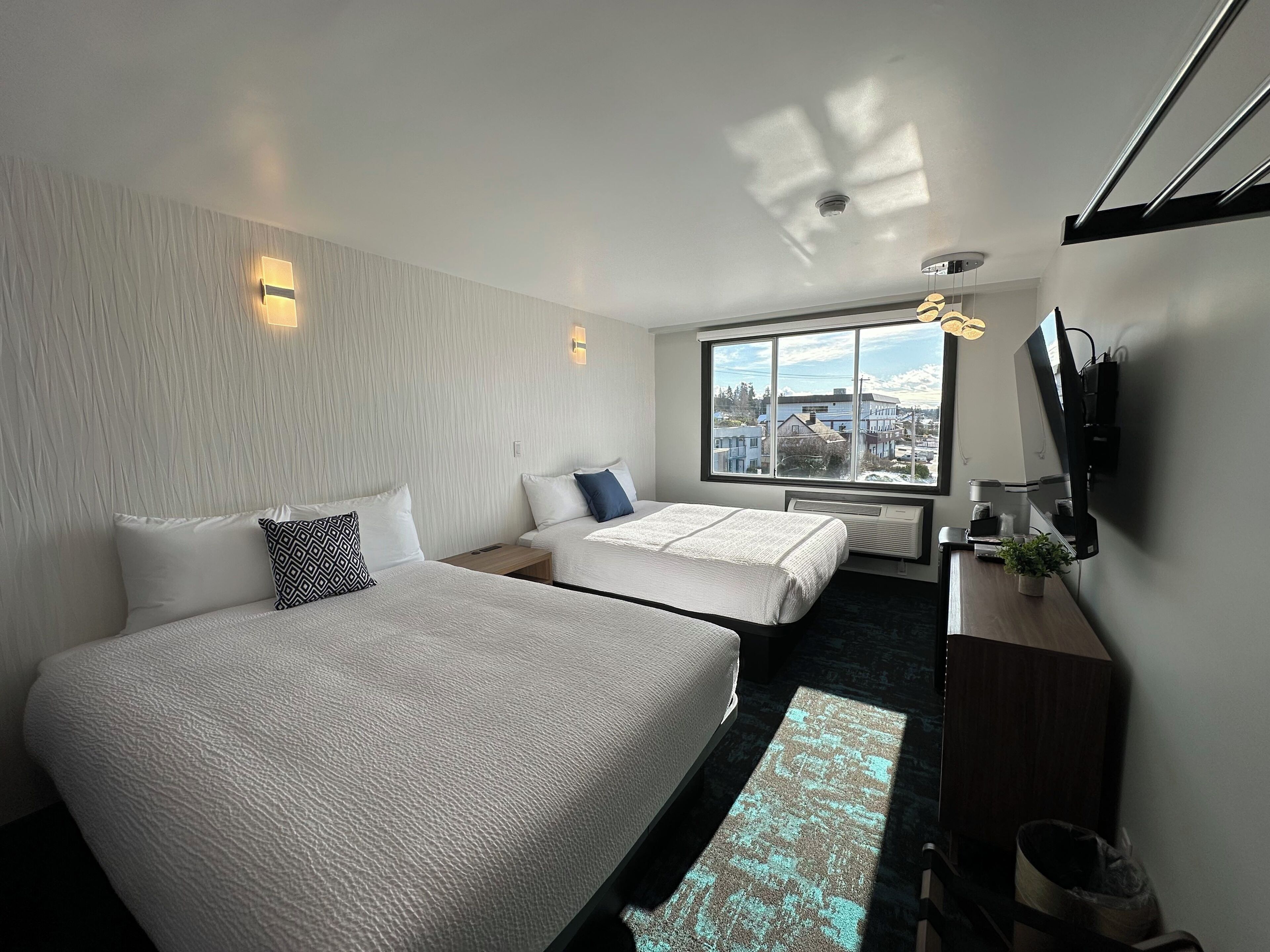 A hotel room with two beds, a large window with a city view, a flat-screen TV mounted on the wall, and a small table with a plant.