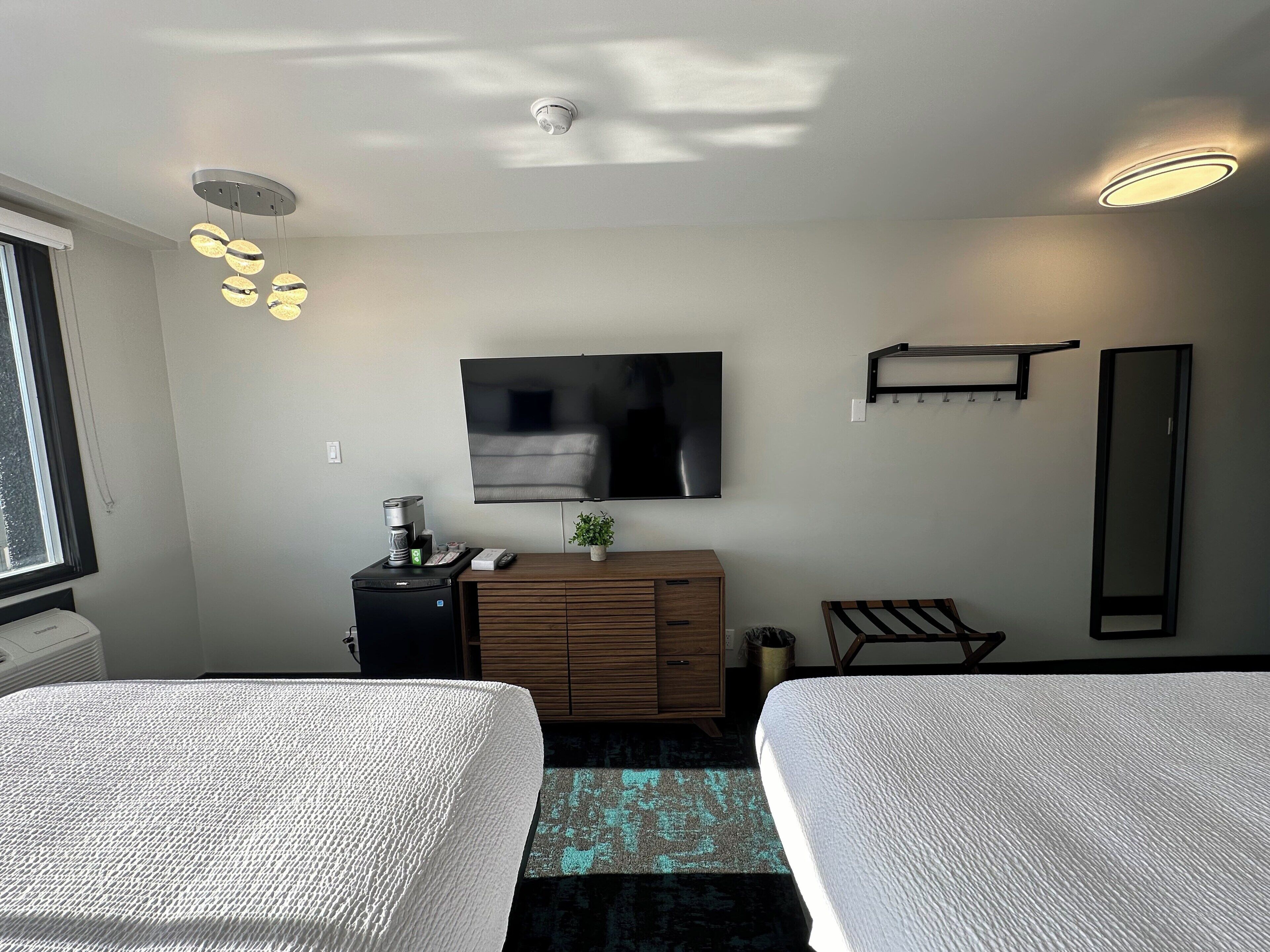 Habitación de hotel con dos camas, un televisor de pantalla plana, un aparador de madera, una mesita con una planta y una ventana con persianas.