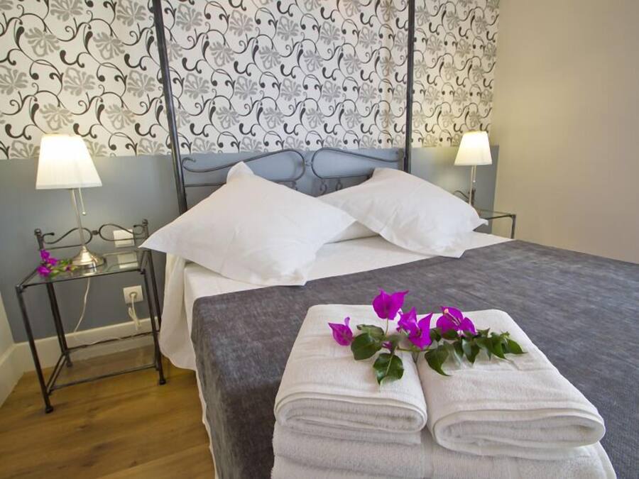 En pænt redt seng med hvide sengetøj og puder, et glasbord ved siden af sengen med en lampe, og et lille bord med en blomst.