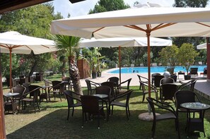 An outdoor seating area with tables and chairs under umbrellas, a pool, and greenery in the background.