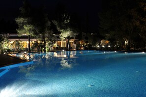 Una piscina nocturna rodeada de árboles y un edificio con ventanas iluminadas.
