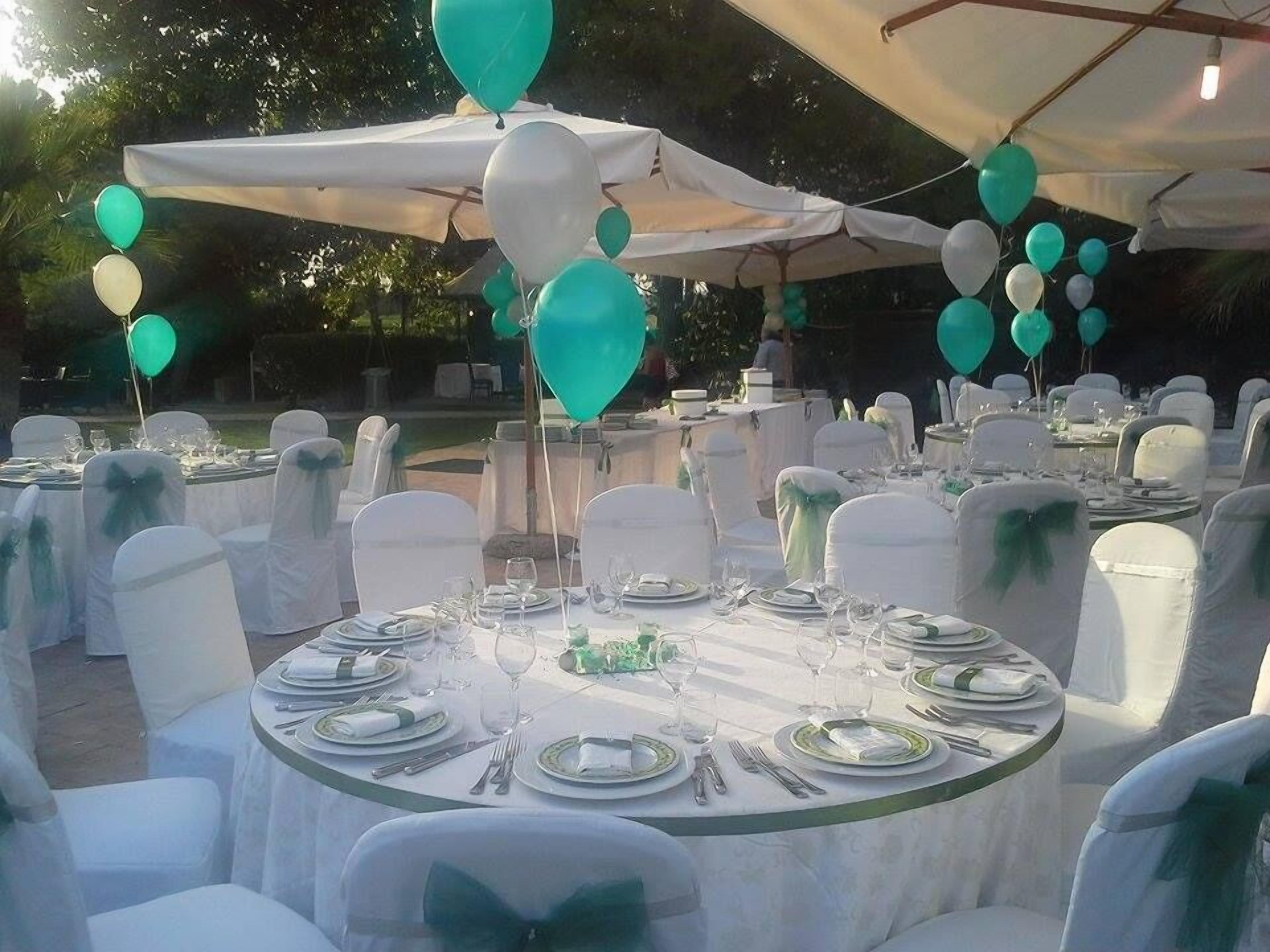 Een evenementenruimte buiten, met ronde tafels voor dineren, witte stoelen met groene linten, en blauwgroene en witte ballonnen.