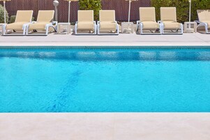 Una piscina con seis sillas reclinables color beige alineadas a lo largo del borde.