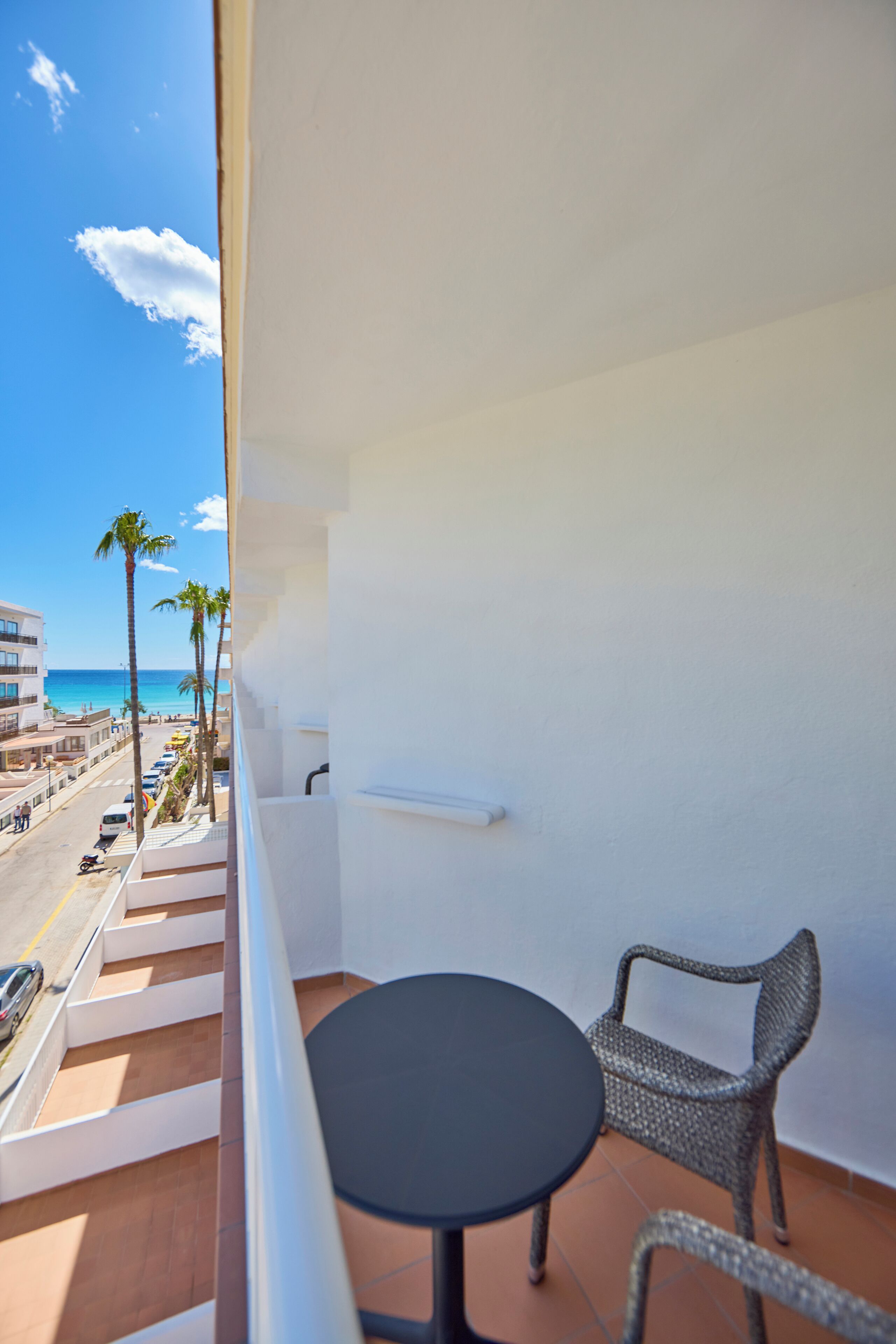 A balcony with a table and chair, overlooking a beach with palm trees and a clear sky.