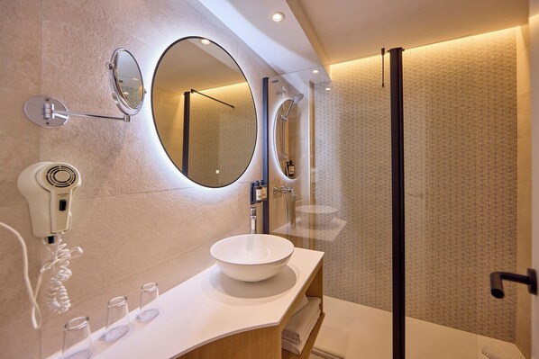 A modern bathroom with a large oval mirror, a round mirror, a white sink, a hairdryer, and a glass shower enclosure. - Universal Hotel Bikini (Sant Llorenc des Cardassar)