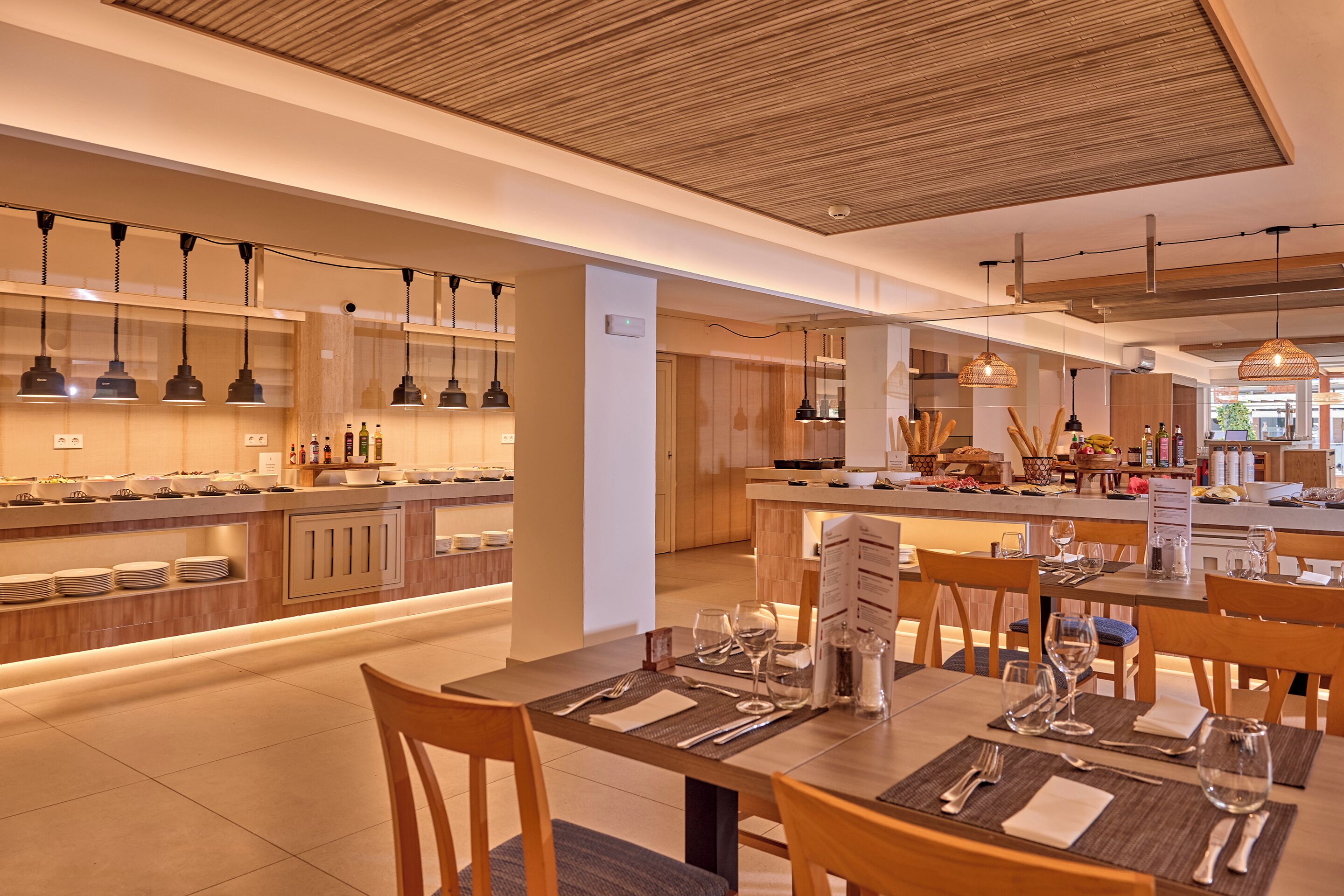 A modern kitchen with a buffet setup, dining area with tables set for two, and a visible ceiling with a wooden design.