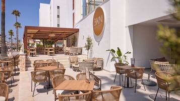 Un espacio al aire libre con mobiliario de mimbre y una sección cubierta, un letrero que dice 'Café & Cocktail Bar', y un cielo azul despejado.