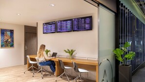A modern office with a wooden desk, two chairs, and a laptop. There are three digital displays on the wall and a potted plant on the right.