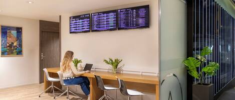 A modern office space with a woman working at a desk, two monitors displaying departure information, and a large window with vertical blinds.
