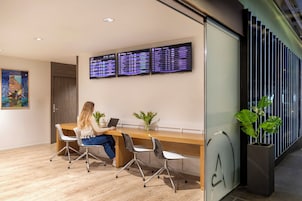 A modern office space with a woman working at a desk, two monitors displaying departure information, and a large window with vertical blinds.