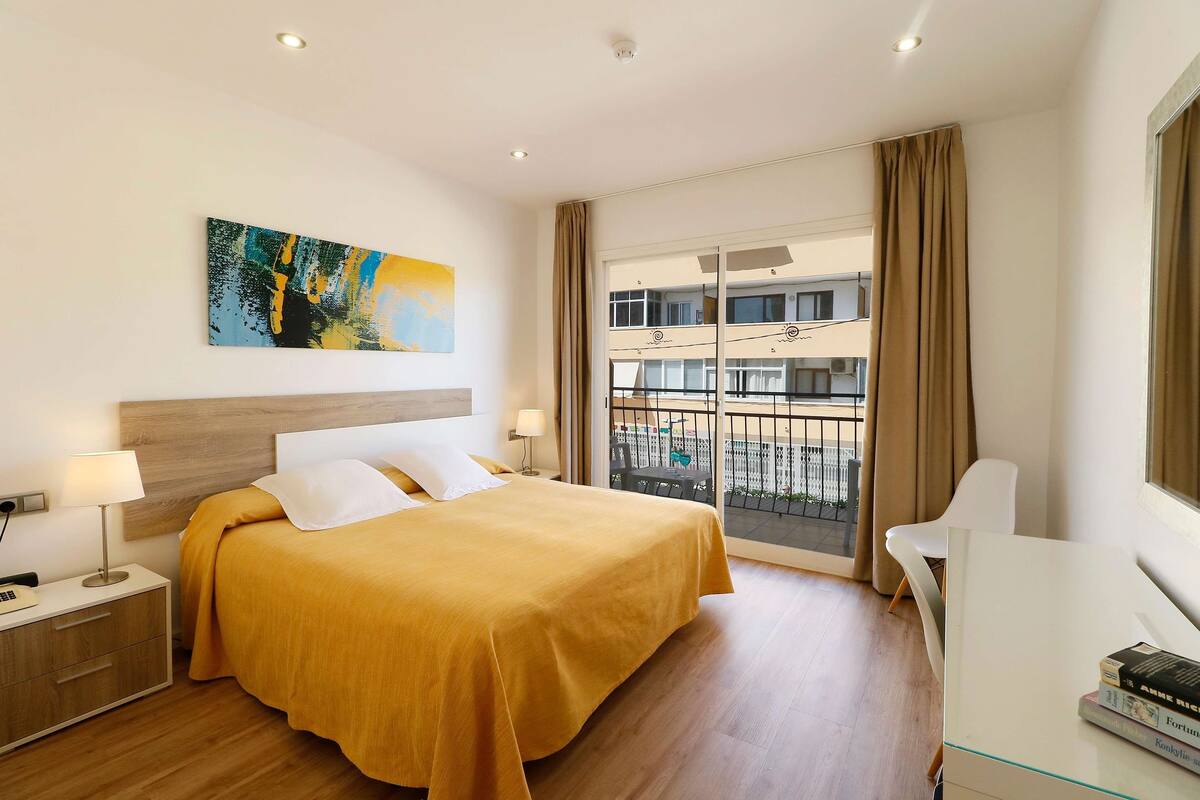 A modern hotel room with a large bed, a desk, a chair, a balcony with a view of the city, and a painting on the wall.