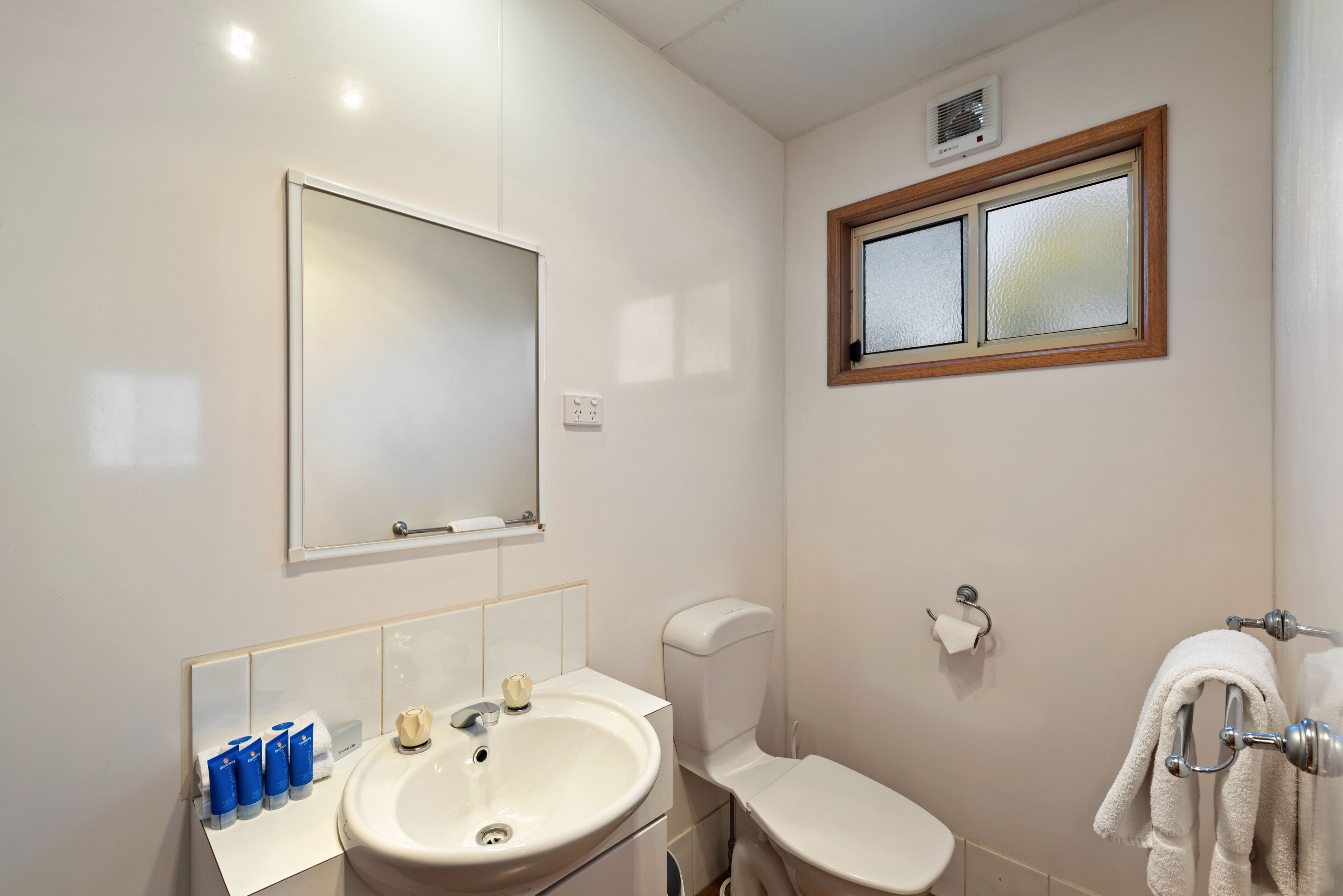 Un baño con lavamanos blanco, espejo, inodoro y ventana.