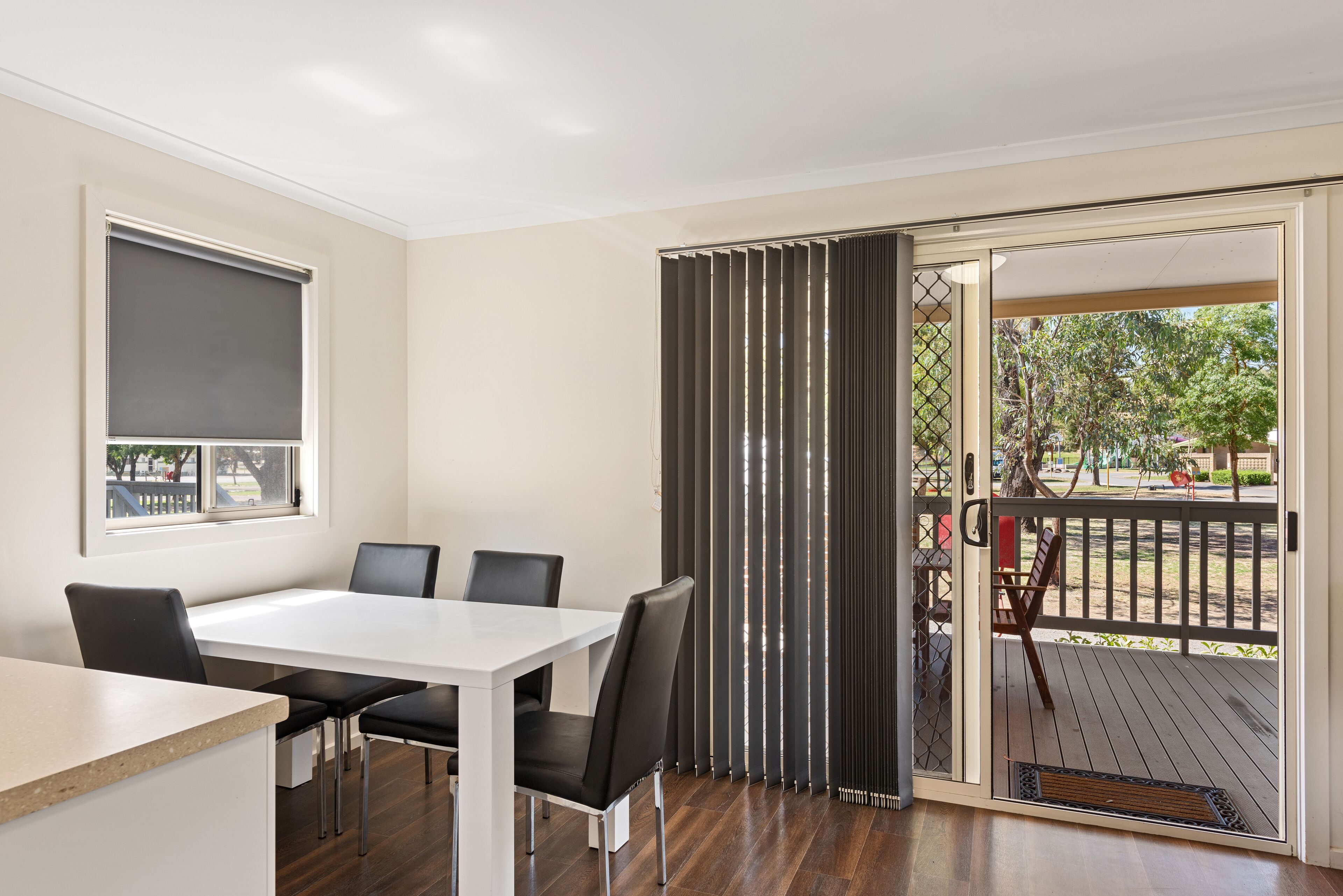 Un coin repas avec une table blanche et des chaises noires, une fenêtre avec un store et une vue sur une terrasse et la verdure à l’extérieur.