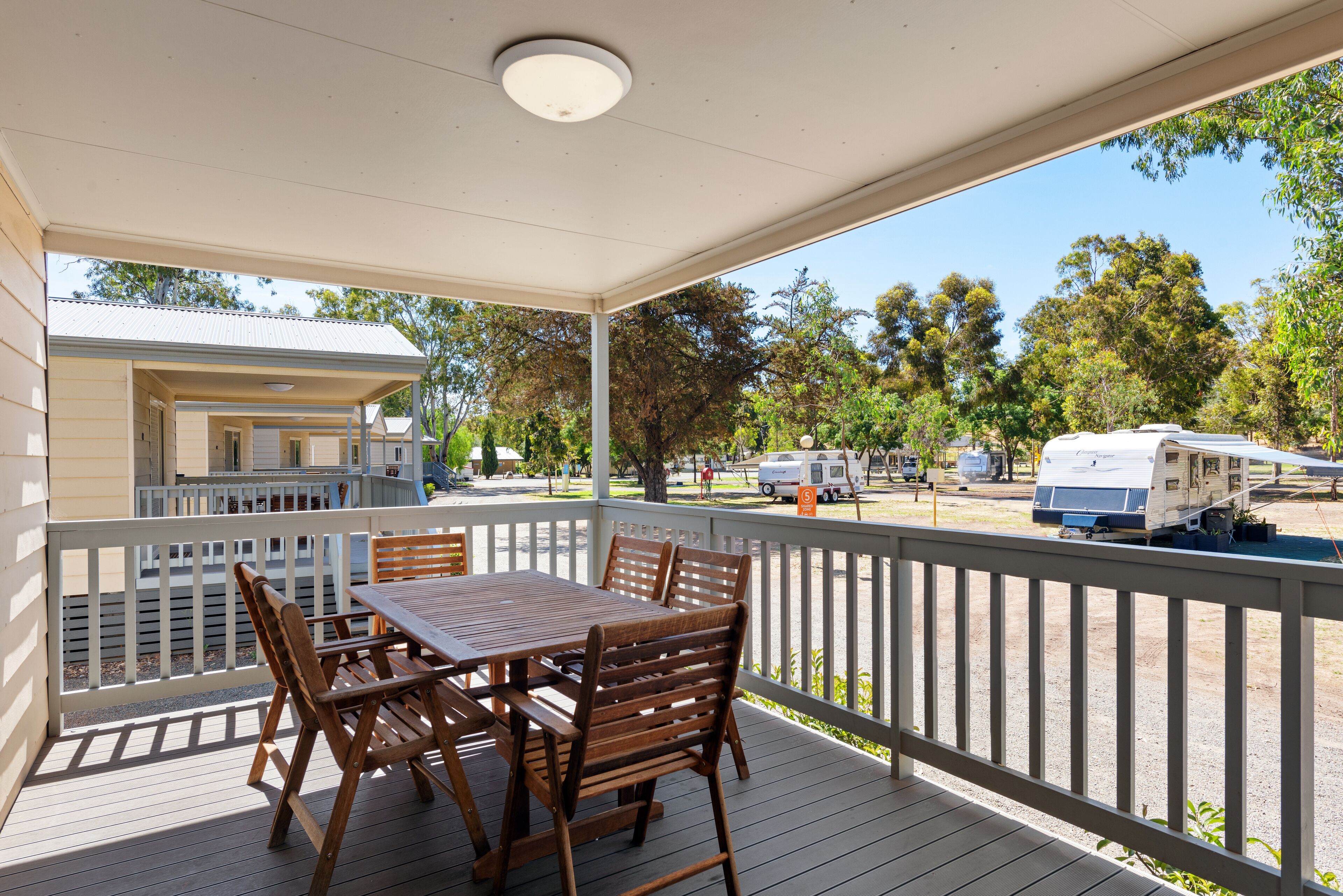 A covered outdoor area with a wooden table and chairs, a white railing, and a view of a campground with RVs.