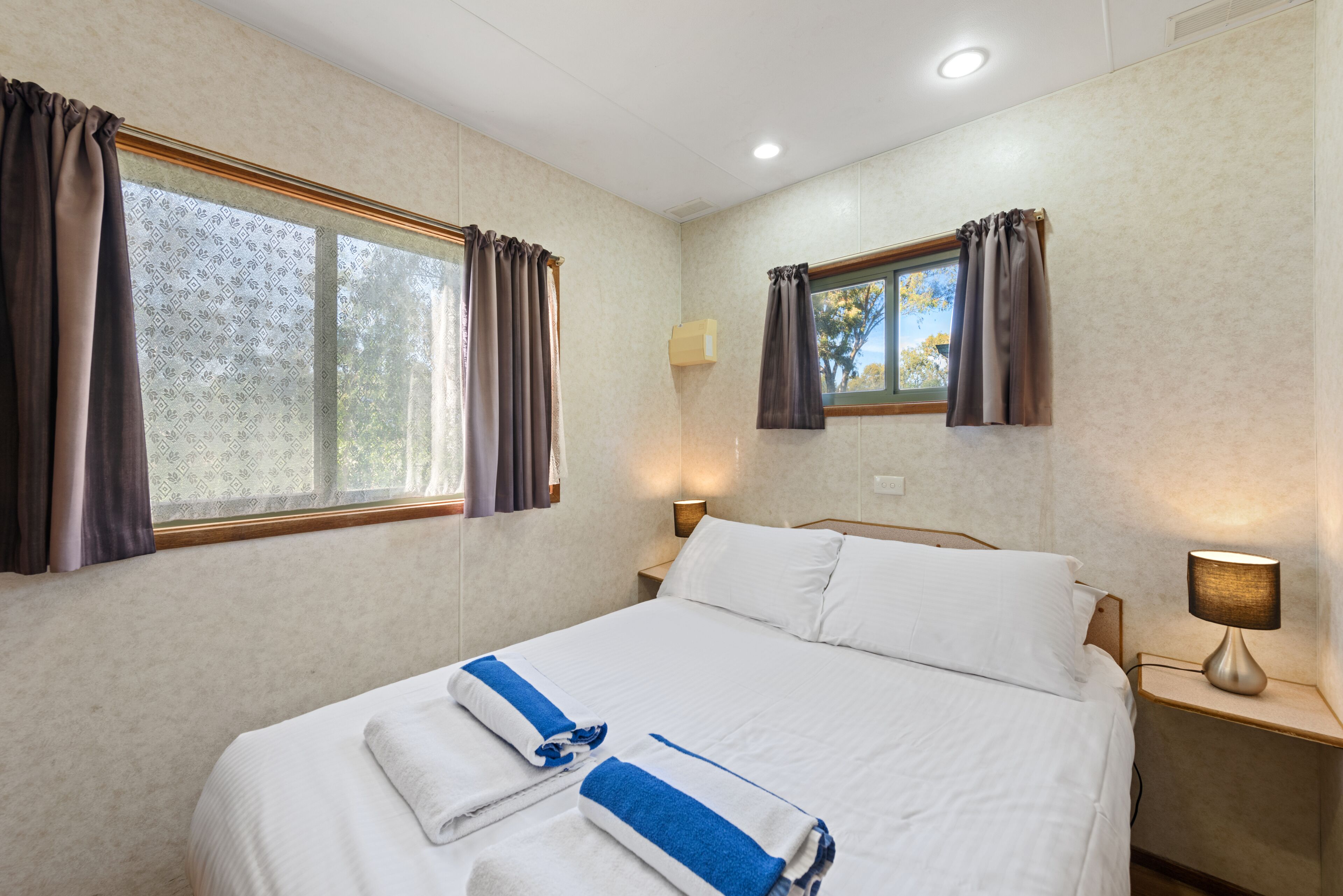 A neatly made bed with white linens and blue towels, two bedside lamps, and a window with curtains.