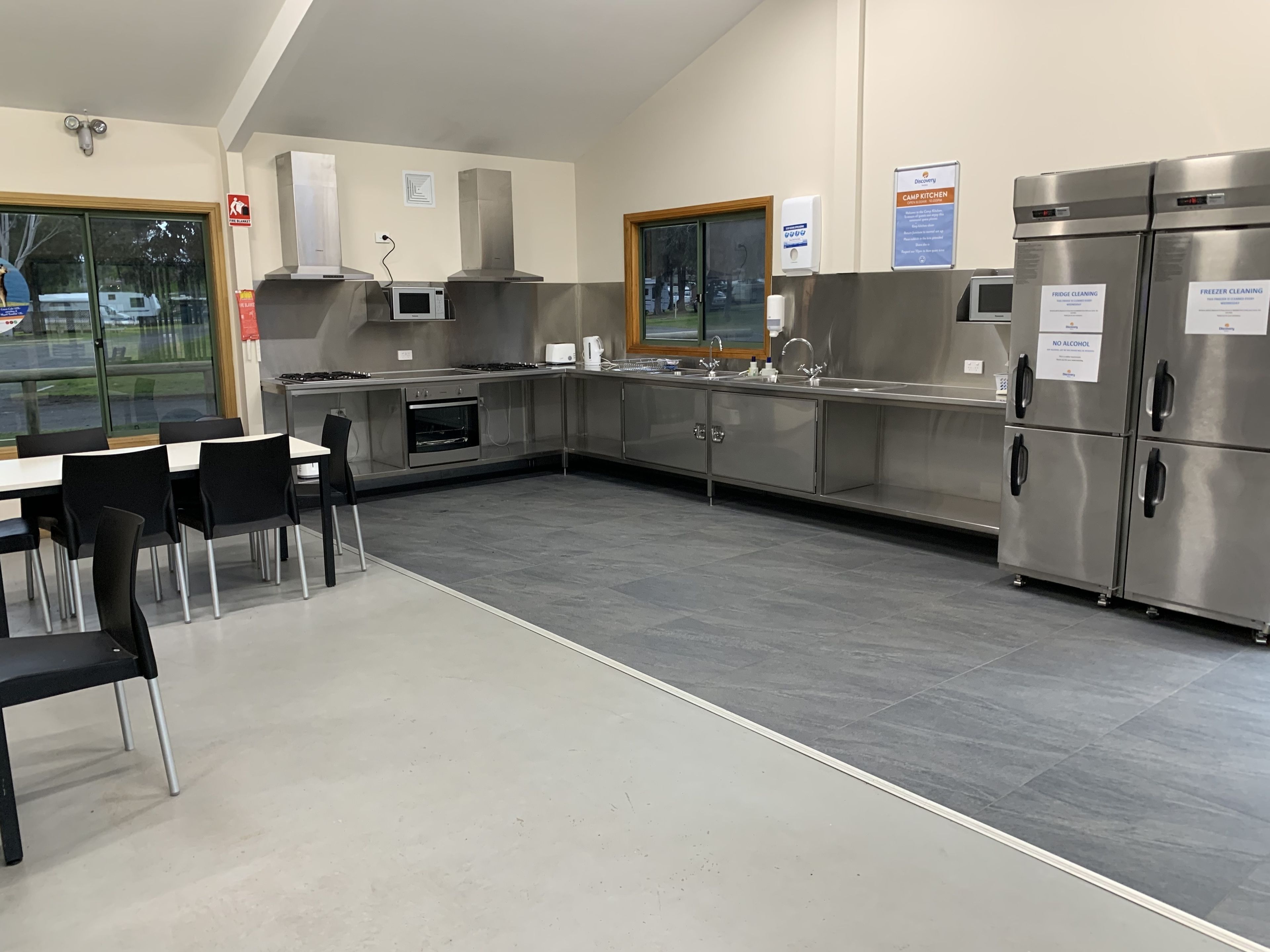 Una cocina moderna con electrodomésticos de acero inoxidable, horno empotrado y un lavamanos. Hay sillas negras y una mesa, y una ventana con vistas al exterior.