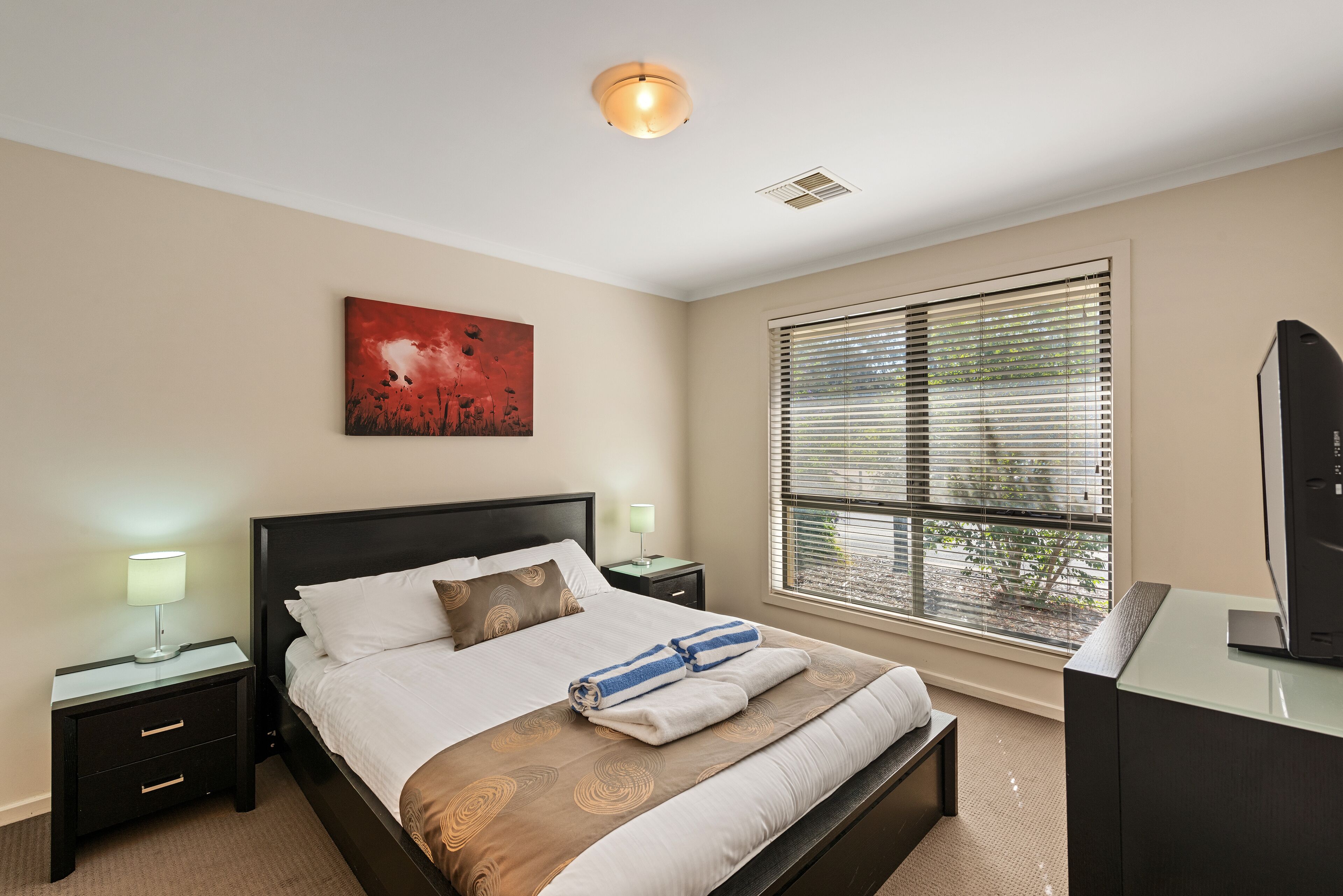 A neatly arranged bedroom with a bed, bedside tables, a painting, a window with blinds, and a television.