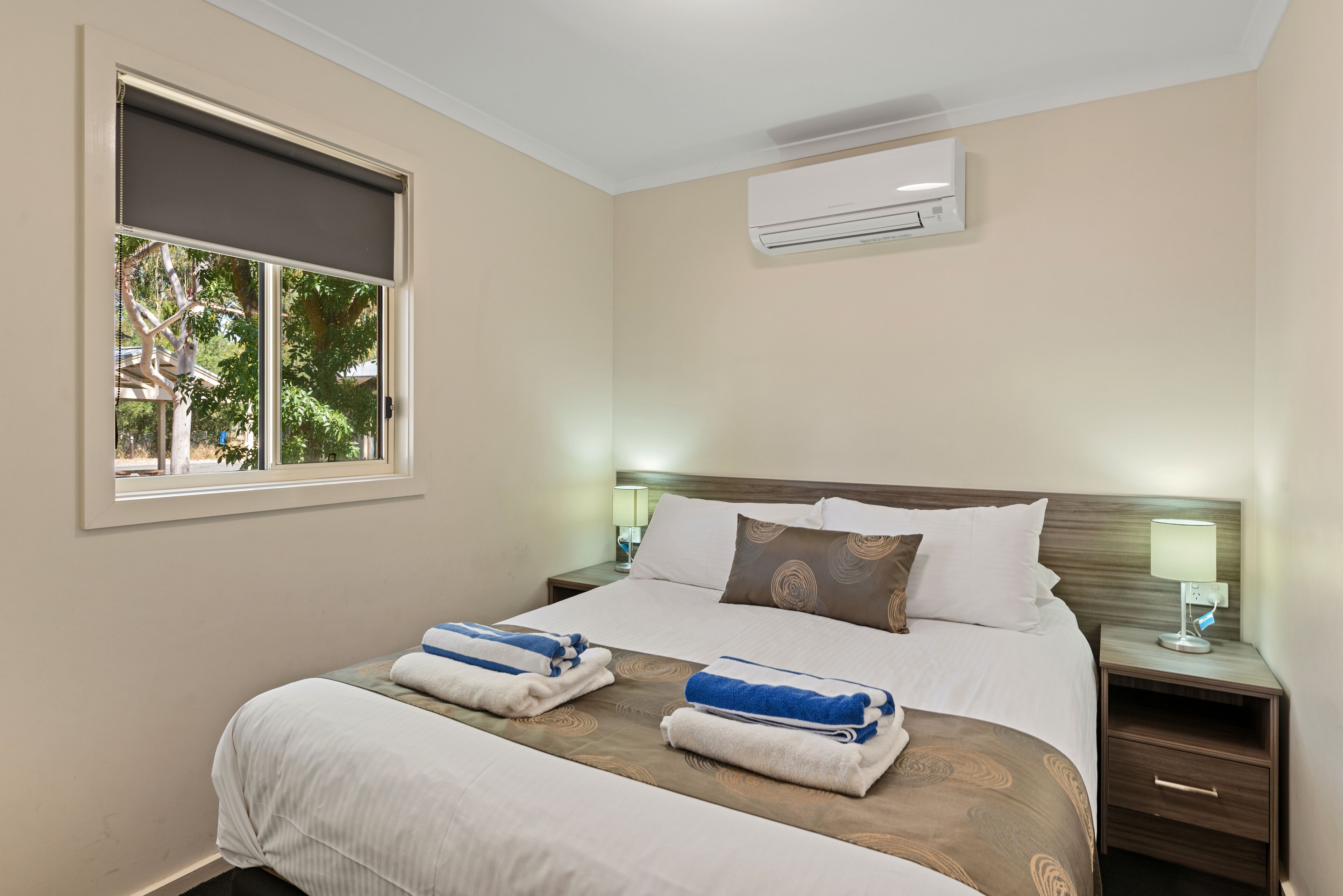 A hotel room with a large bed, two bedside tables, a window, and an air conditioning unit.