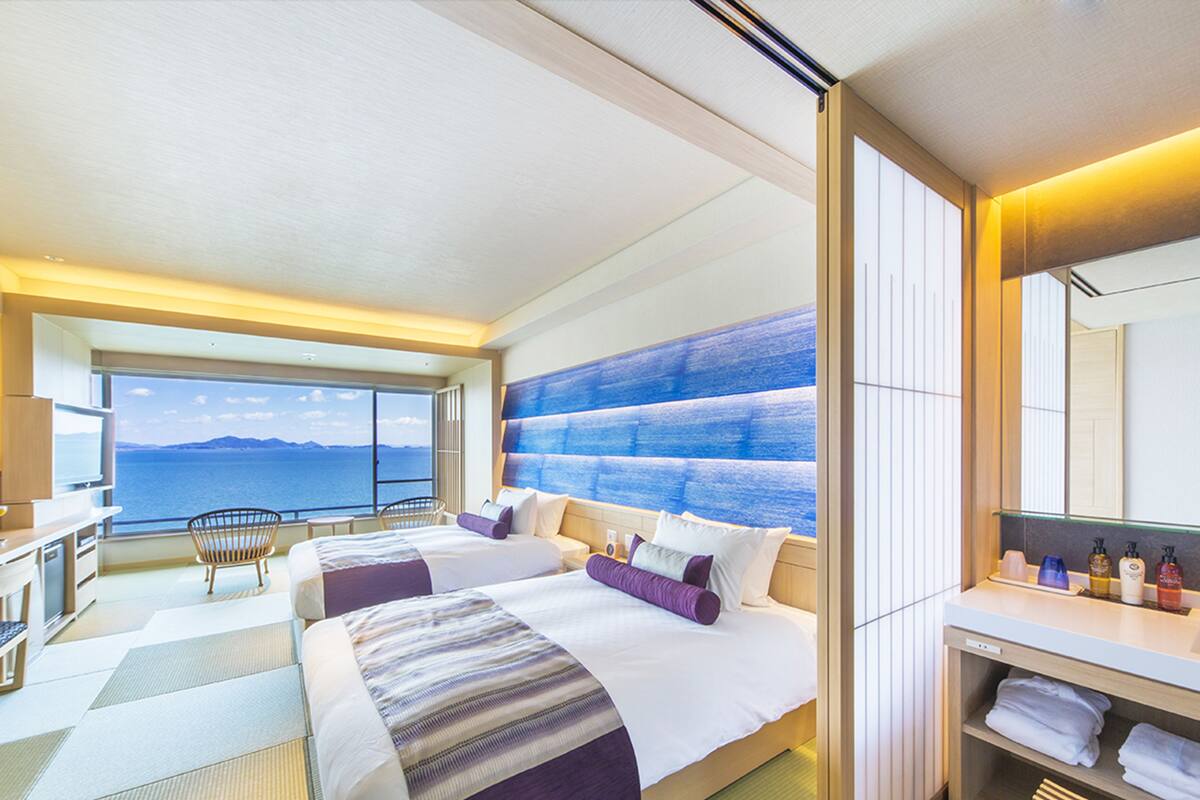 A hotel room with two beds, a large window with a view of the sea, a desk with a chair, and a vanity with toiletries.