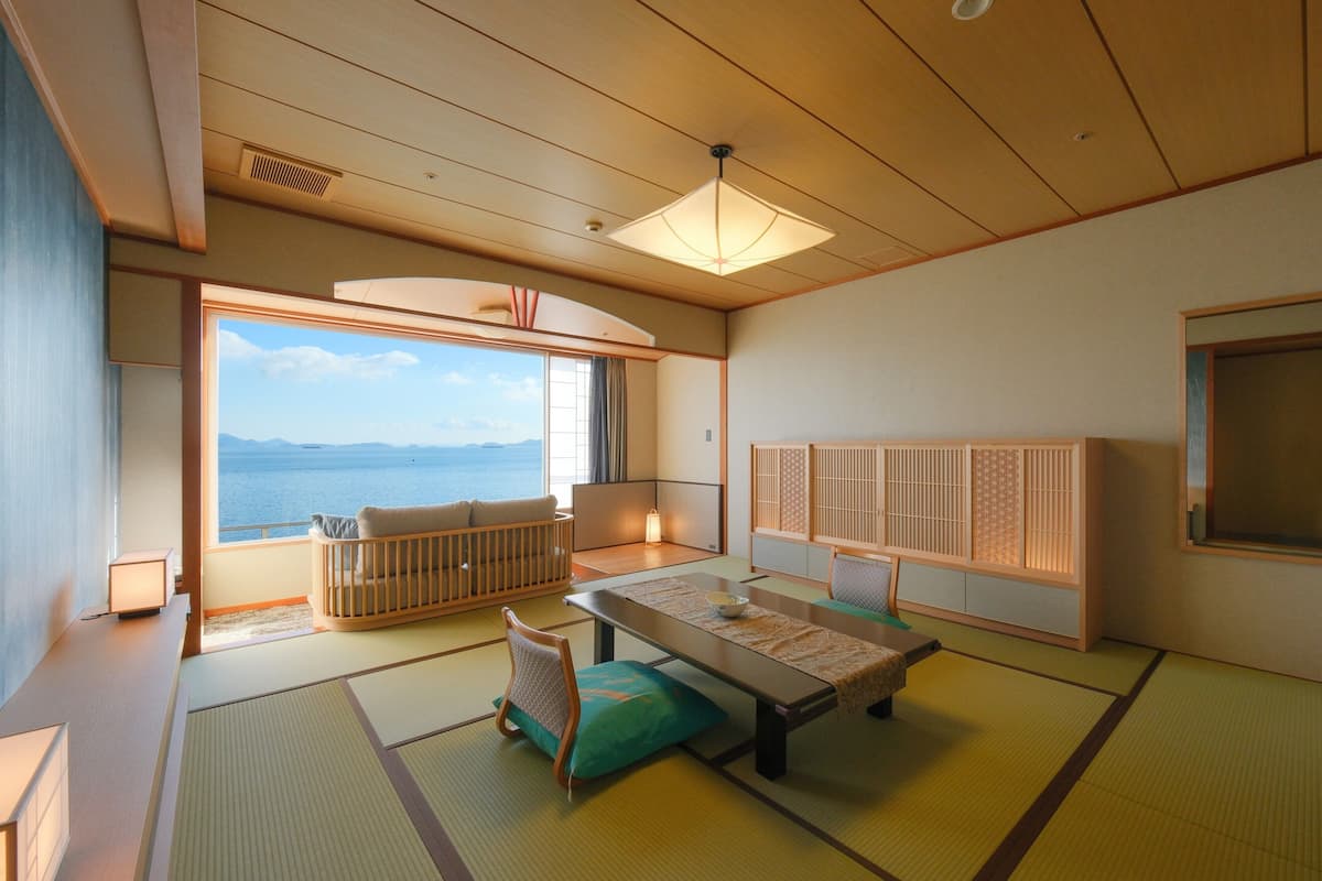 A traditional Japanese room with tatami flooring, a low table, cushions, and a sliding door leading to a balcony with a view of the sea.