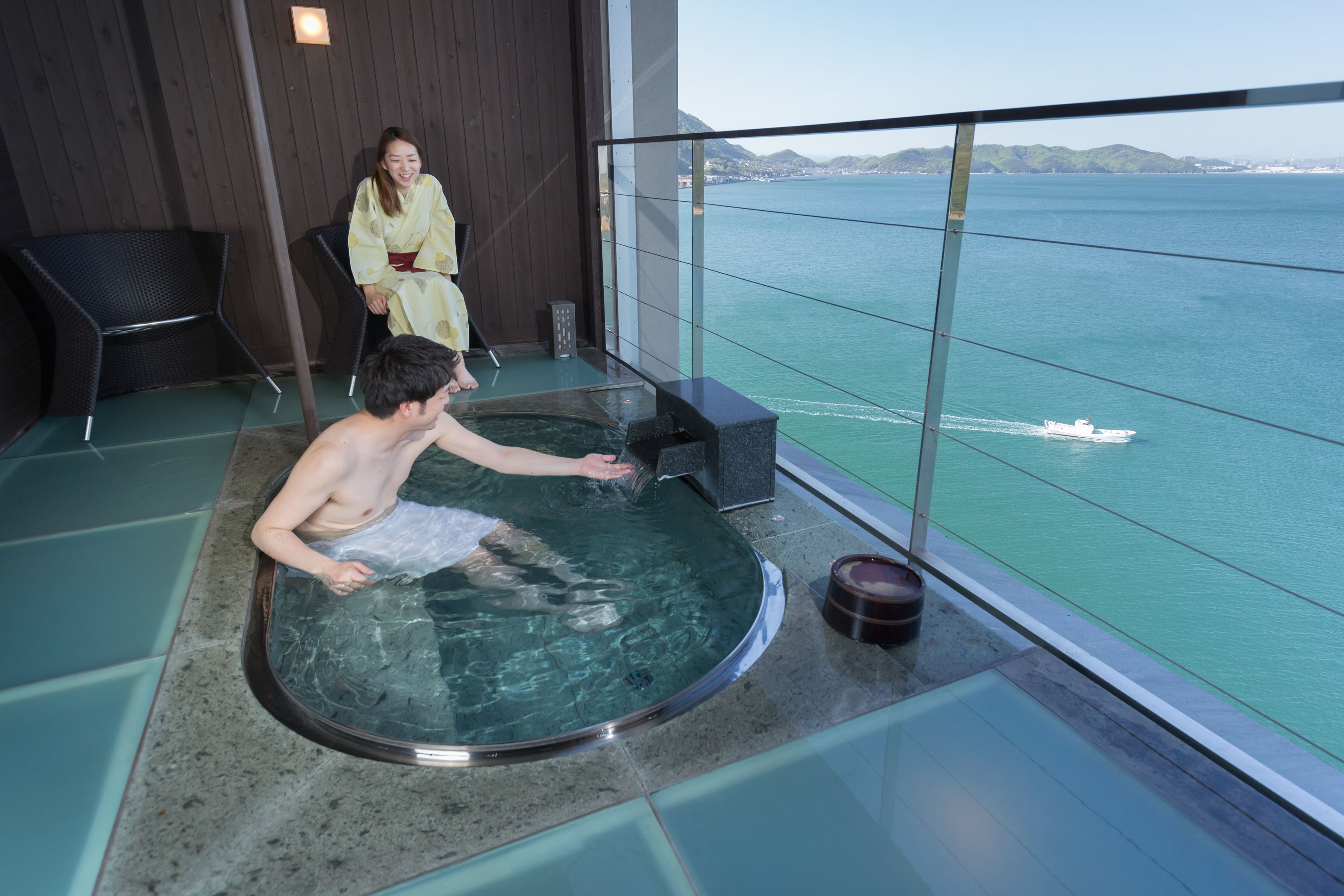 A man in a hot tub on a balcony with a view of the sea.