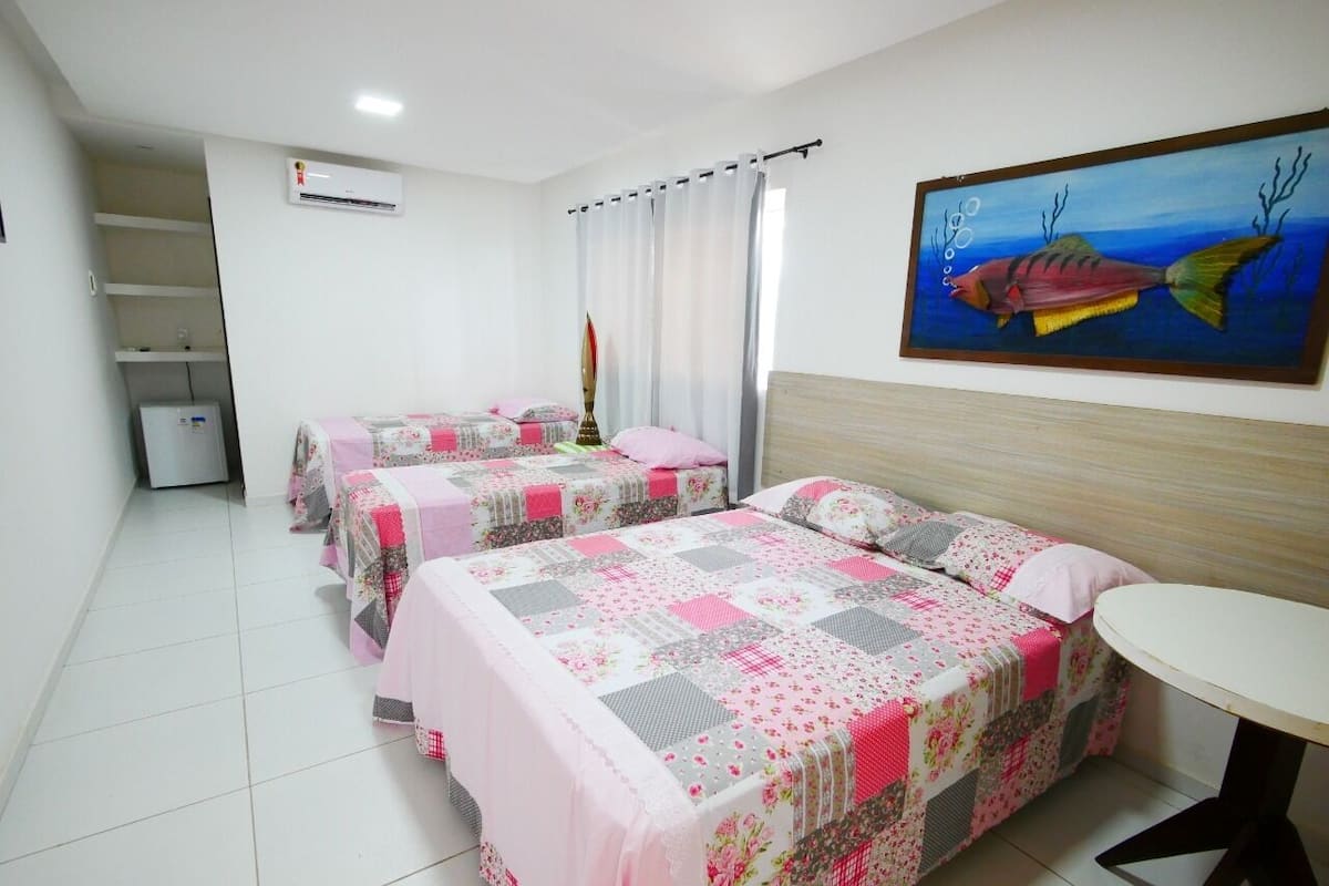 Une chambre d’hôtel avec trois lits, une petite table, un tableau au mur et un réfrigérateur.
