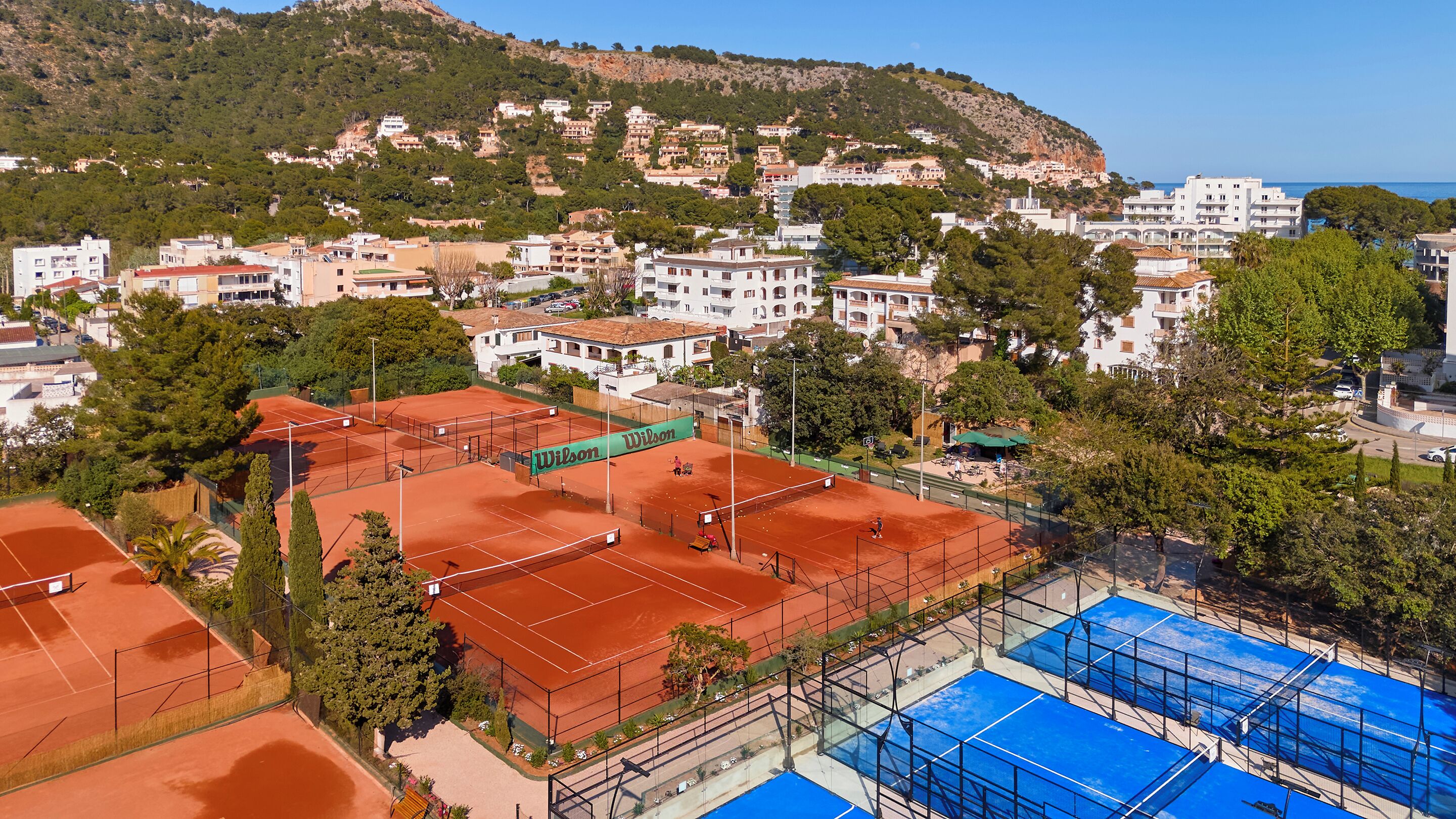 Een tenniscomplex met meerdere banen, omgeven door woongebouwen en een glooiend landschap op de achtergrond.