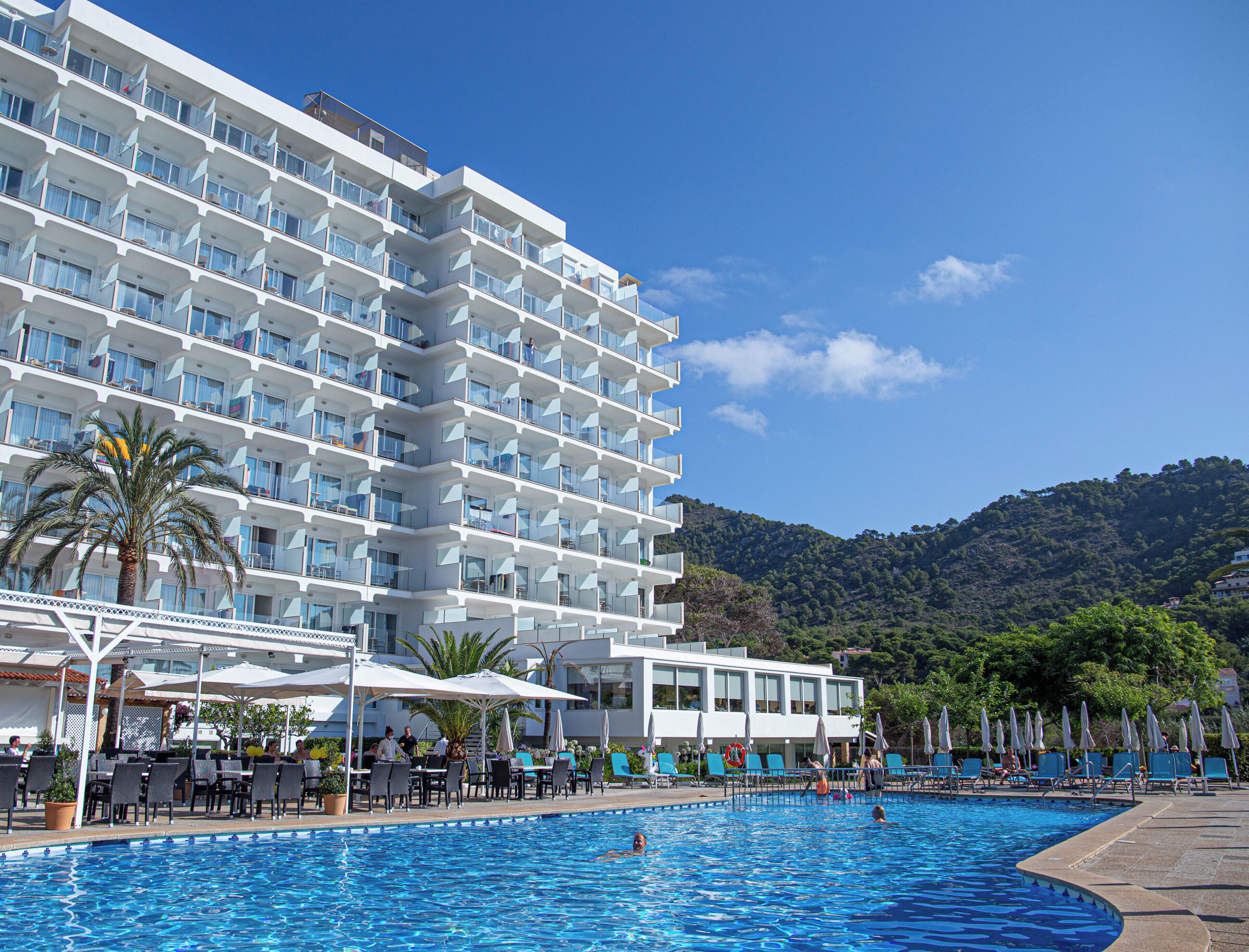 Ein Hotel mit mehreren Balkonen, einem Swimmingpool, Sitzgelegenheiten im Freien und Blick auf einen Hügel.