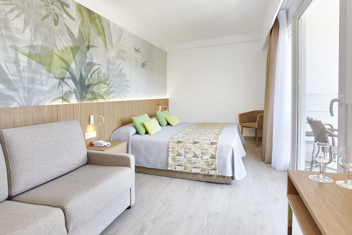 A modern hotel room with a bed, a sofa, a wooden nightstand, and a large window with curtains.