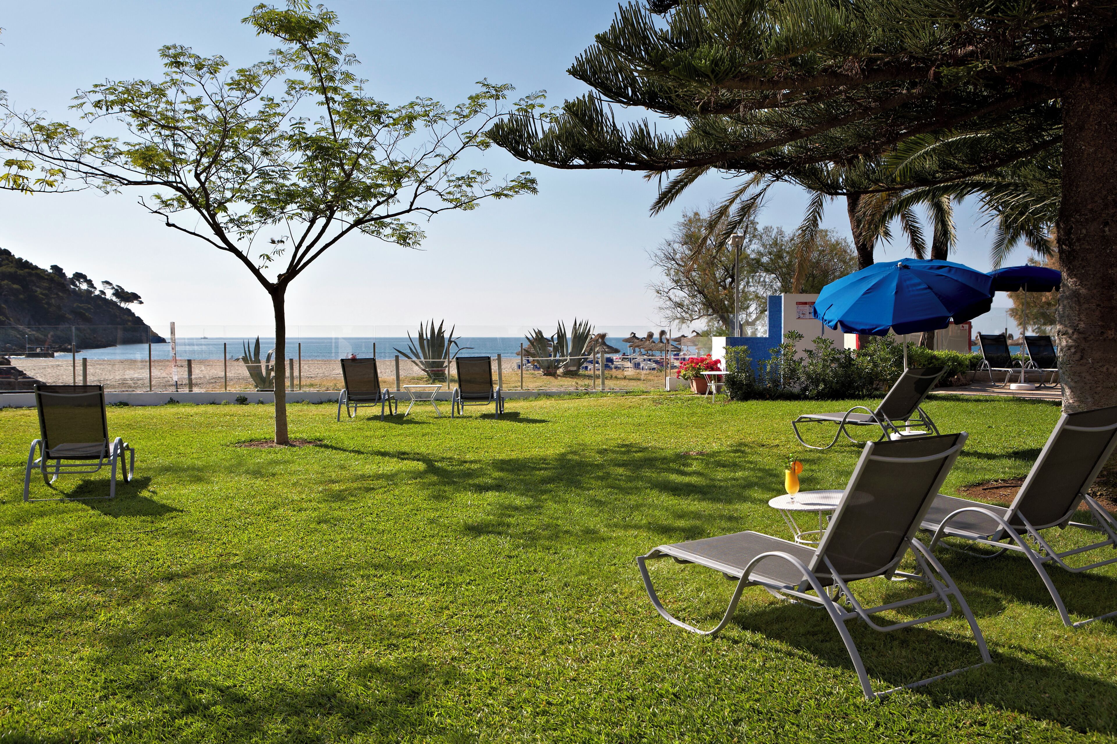 Zona verde al aire libre con sillones de descanso y una sombrilla azul, con vista a la playa.