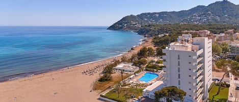 A coastal hotel with a pool, beach access, and a view of the sea and hills.